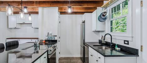A modern kitchen with white cabinetry, a black countertop, and a wooden ceiling. There are hanging lights, a window with a view, and a sink with a faucet.