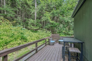 A wooden deck with outdoor furniture, surrounded by dense forest. - Cozy Quechee Hollow Condo with Pond Views & Club Access (Hartford)