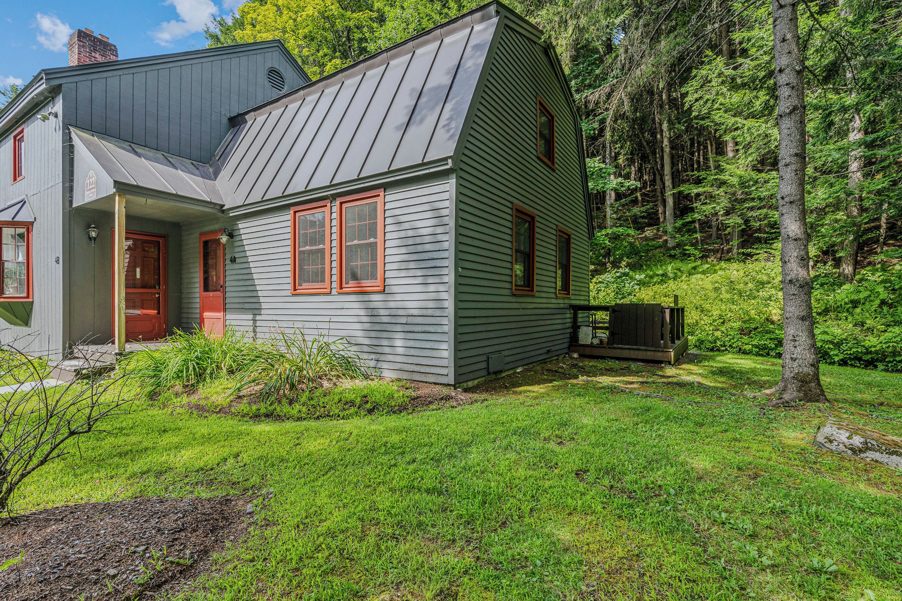 A two-story house with a grey exterior and red trim, surrounded by a lush green landscape and trees.