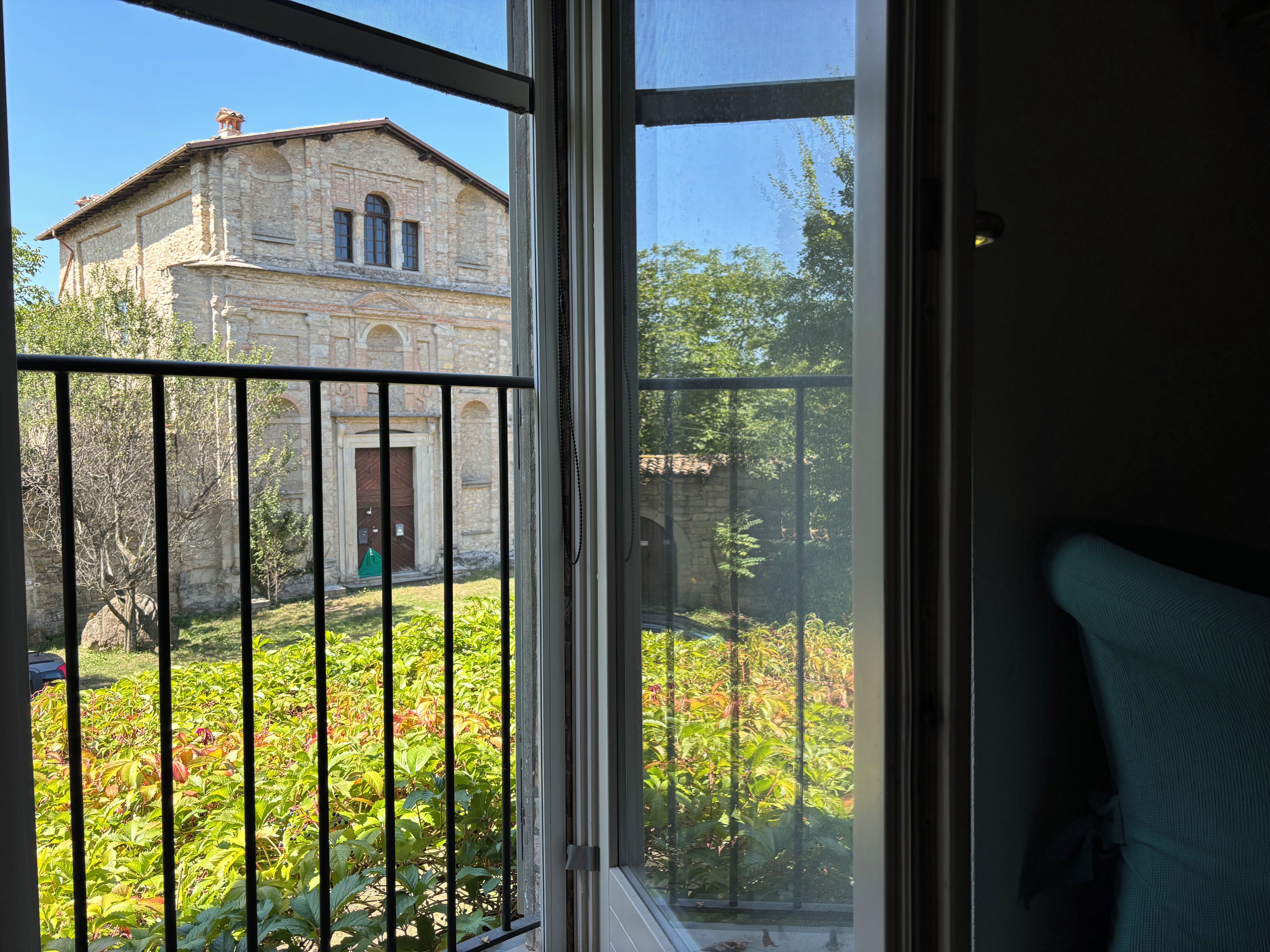A view through a window with a metal railing, a stone building, and a garden with various plants.