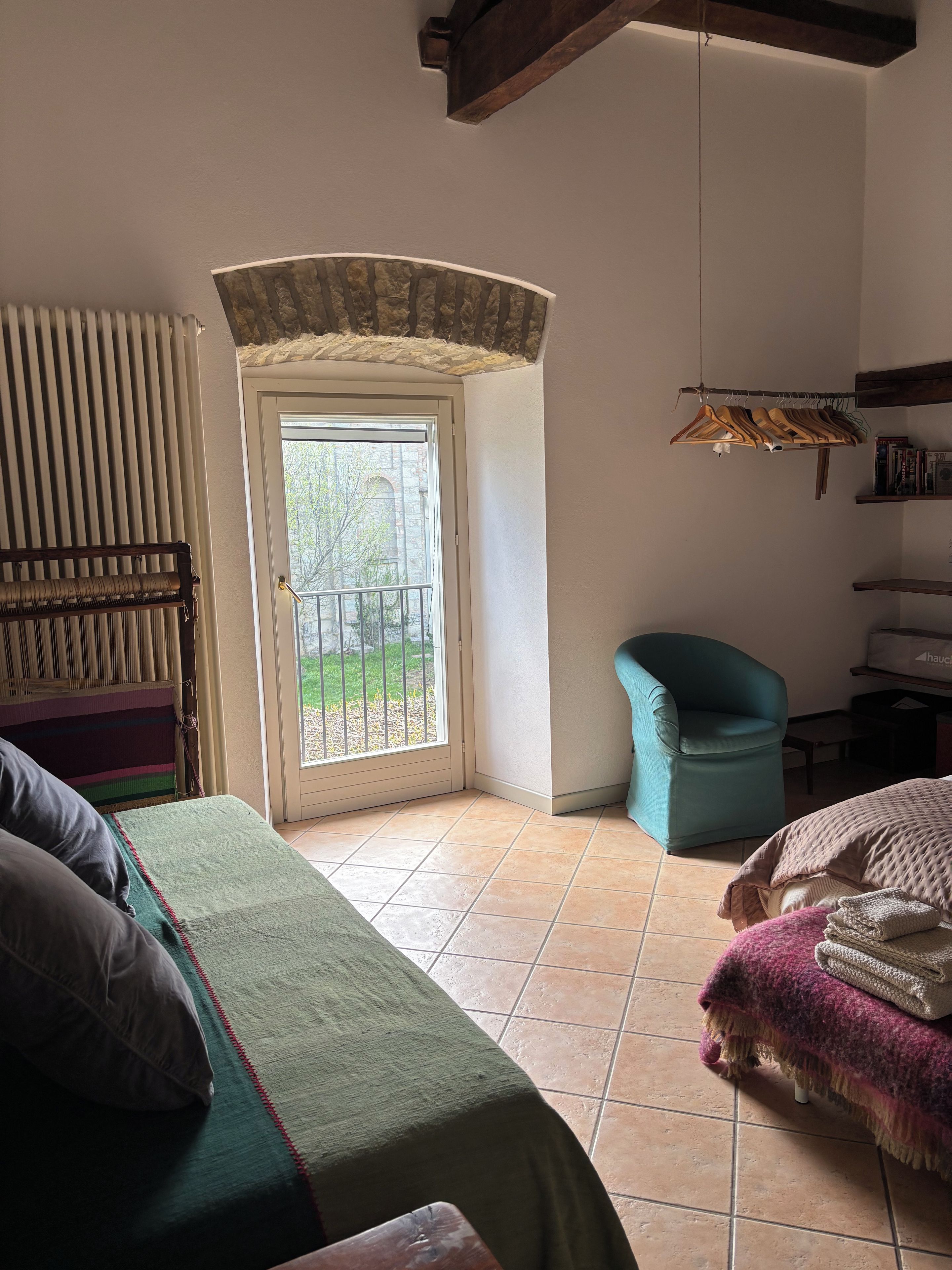 A room with a bed, a chair, a radiator, a door leading to a balcony, and a shelf with books.