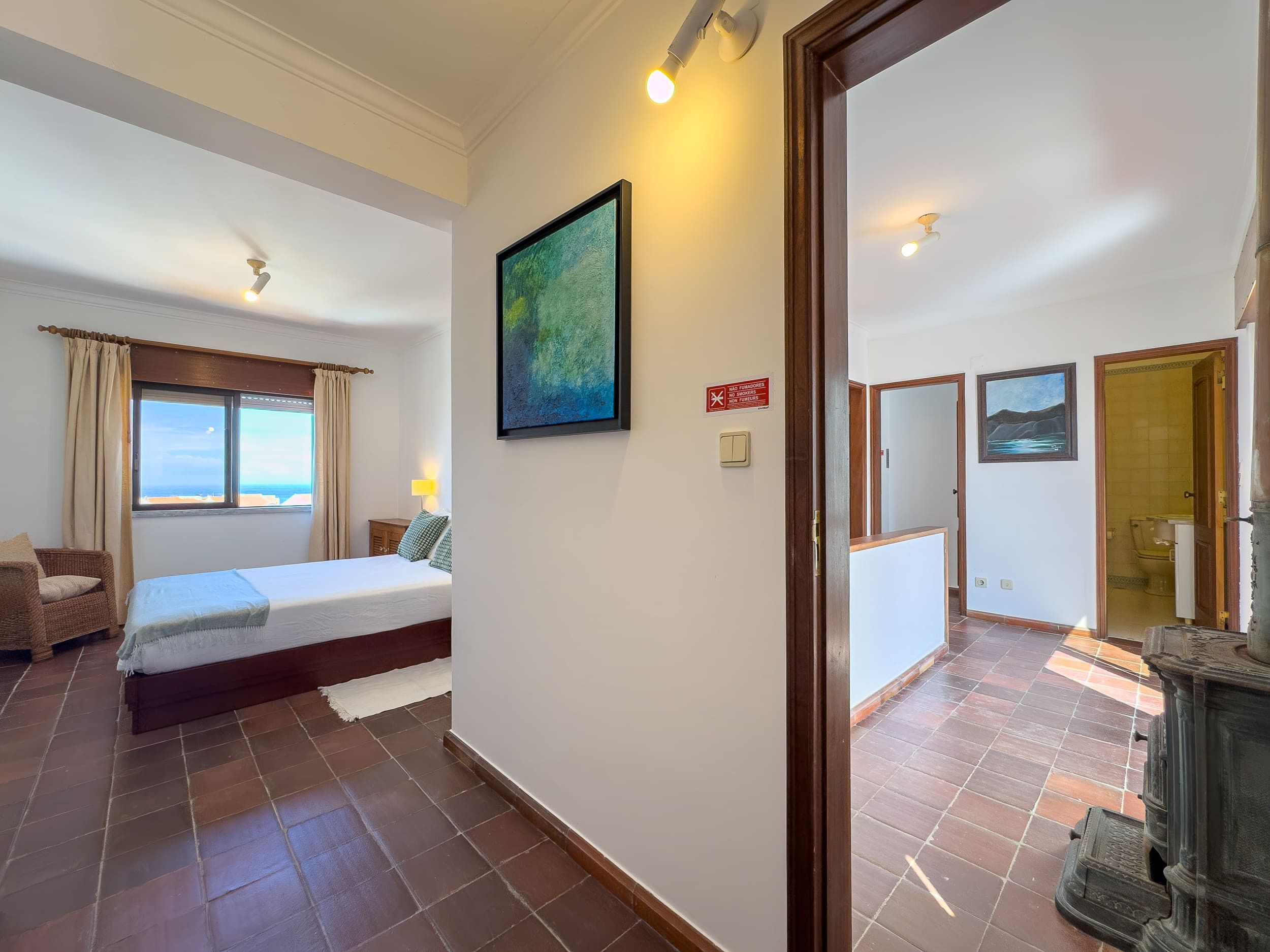 A hotel room with a bed, a view of the sea, a painting on the wall, and a wooden door leading to another room.