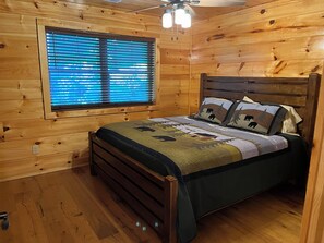 Ein Schlafzimmer einer Holzhütte mit einem Bett, einem Fenster mit Jalousien und einem Deckenventilator.