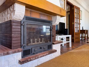 A stone fireplace with a glass door, flanked by brick and stone walls, in a room with a wooden door and a white table. - Alto da Villa by Acasadascasas (Mafra)