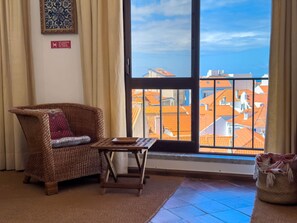 A room with a wicker chair, a small table, a view of buildings through a window, and a sign on the wall. - Alto da Villa by Acasadascasas (Mafra)