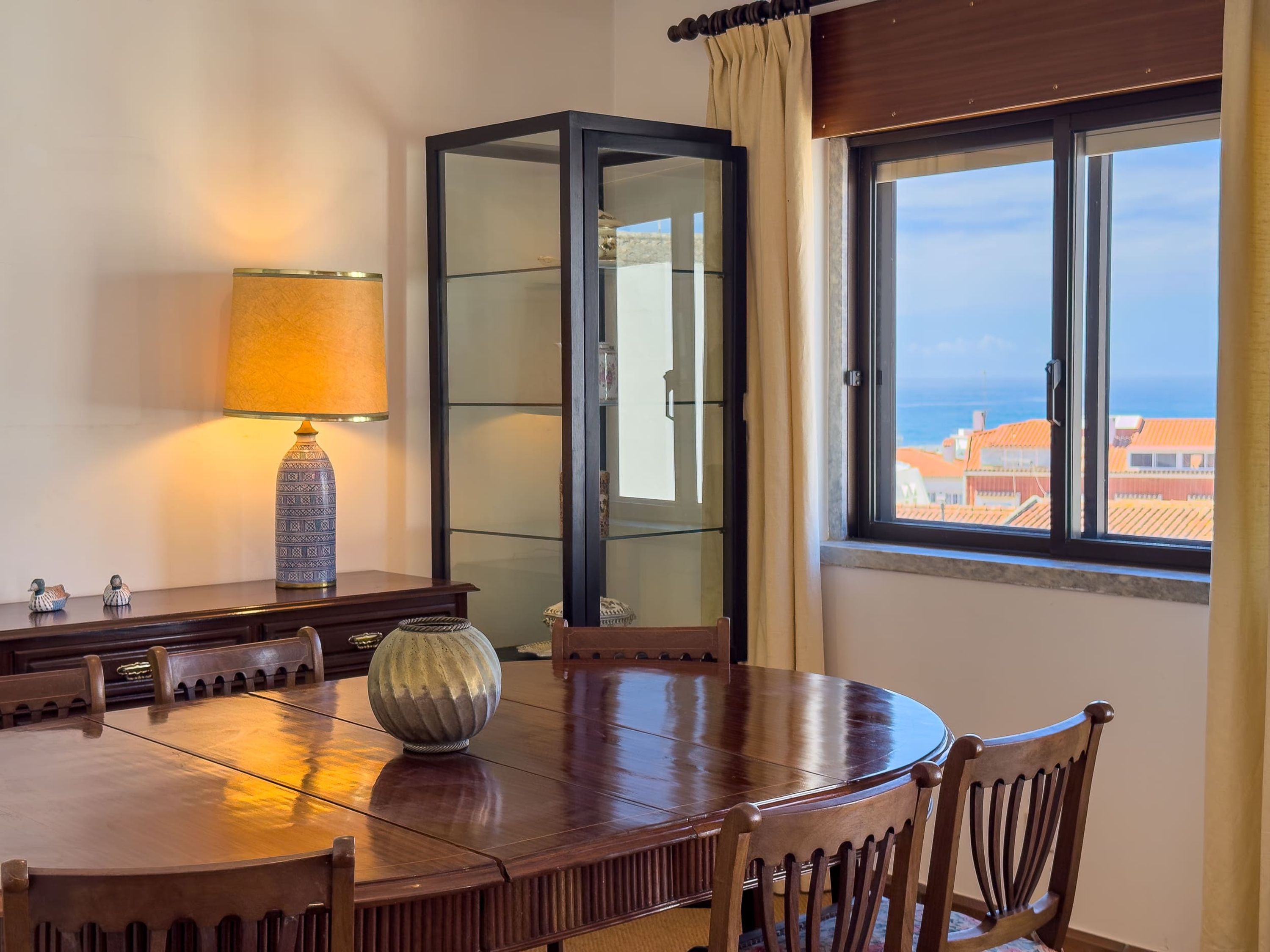 A dining area with a round table, chairs, a lamp, a cabinet, and a window with a view.