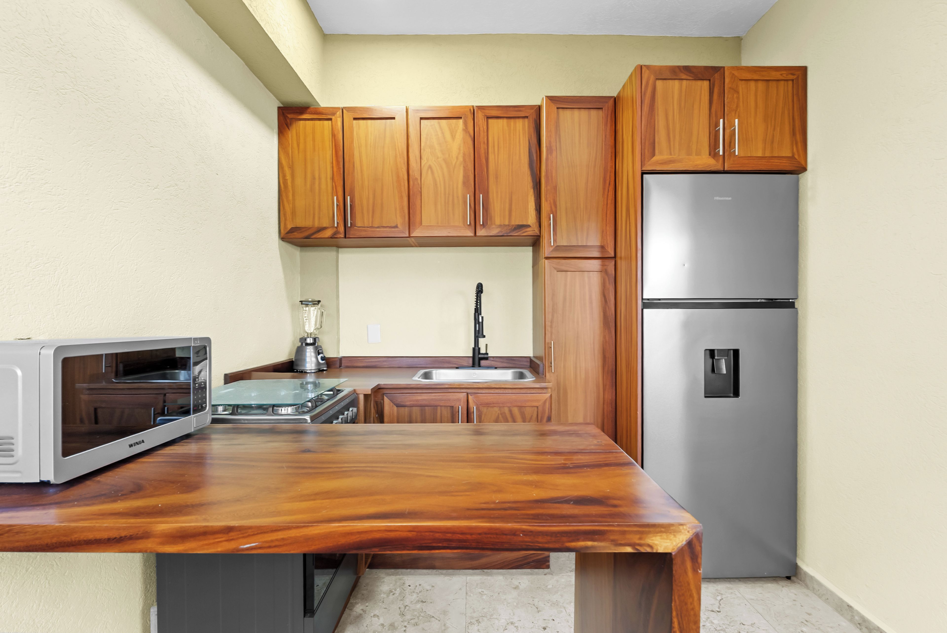 A compact kitchen with wooden cabinets, a stainless steel refrigerator, a microwave, and a blender on the counter.