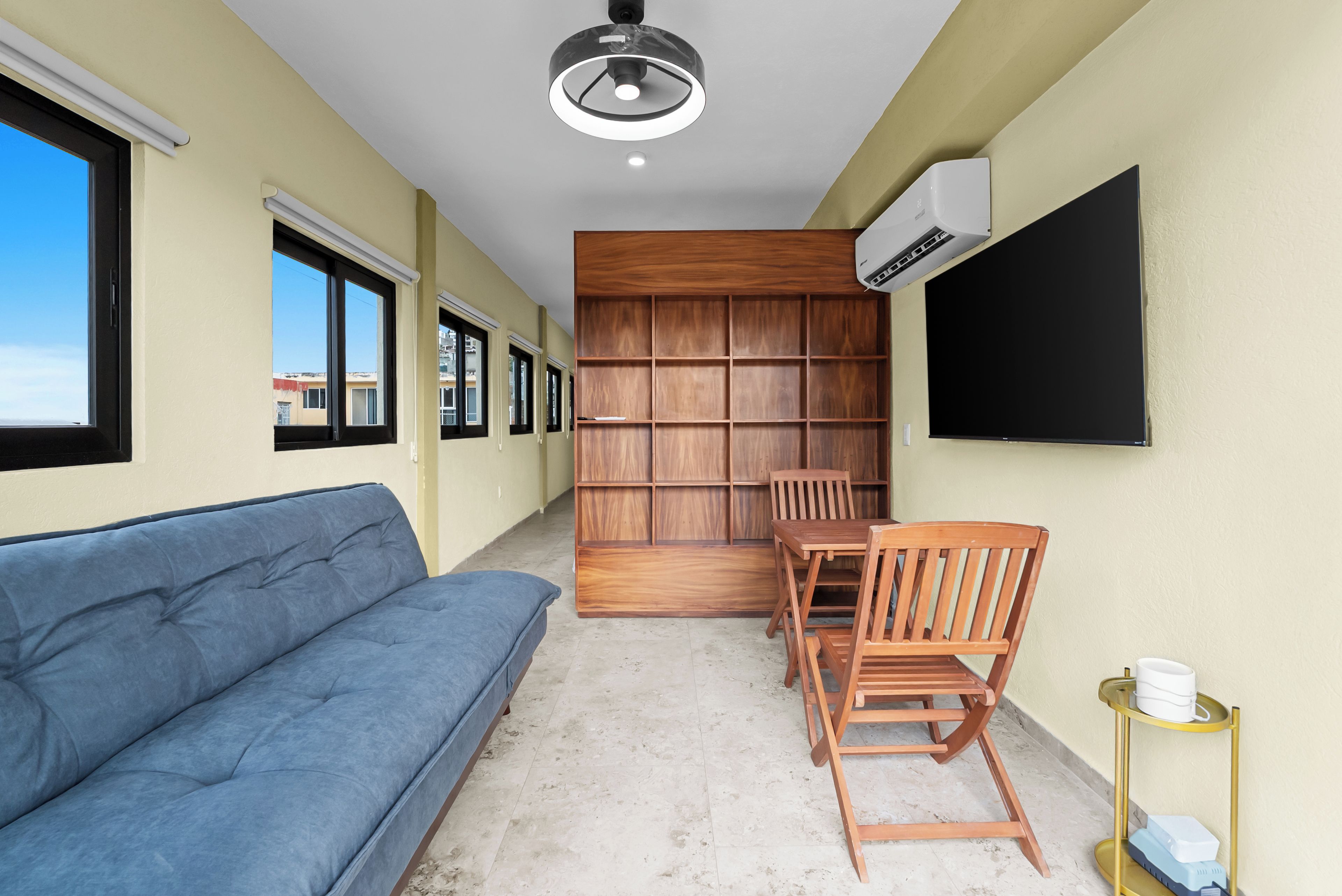 A room with a blue sofa, a wooden bookshelf, a table with chairs, and a mounted TV.