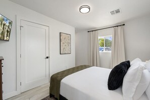 A bedroom with a bed, a door, a window with curtains, and a wall-mounted picture. - Phoenix Furnished with 2 Queen Beds & EV Charger! (Phoenix)