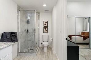 Uma casa de banho moderna com cabine de duche em vidro, sanita e cama visível através de uma porta de vidro.