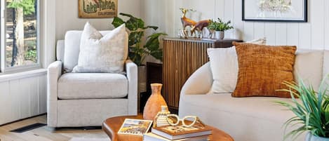 Una sala de estar con un sillón gris, un sofá beige y una mesa de centro de madera con libros y vasos.