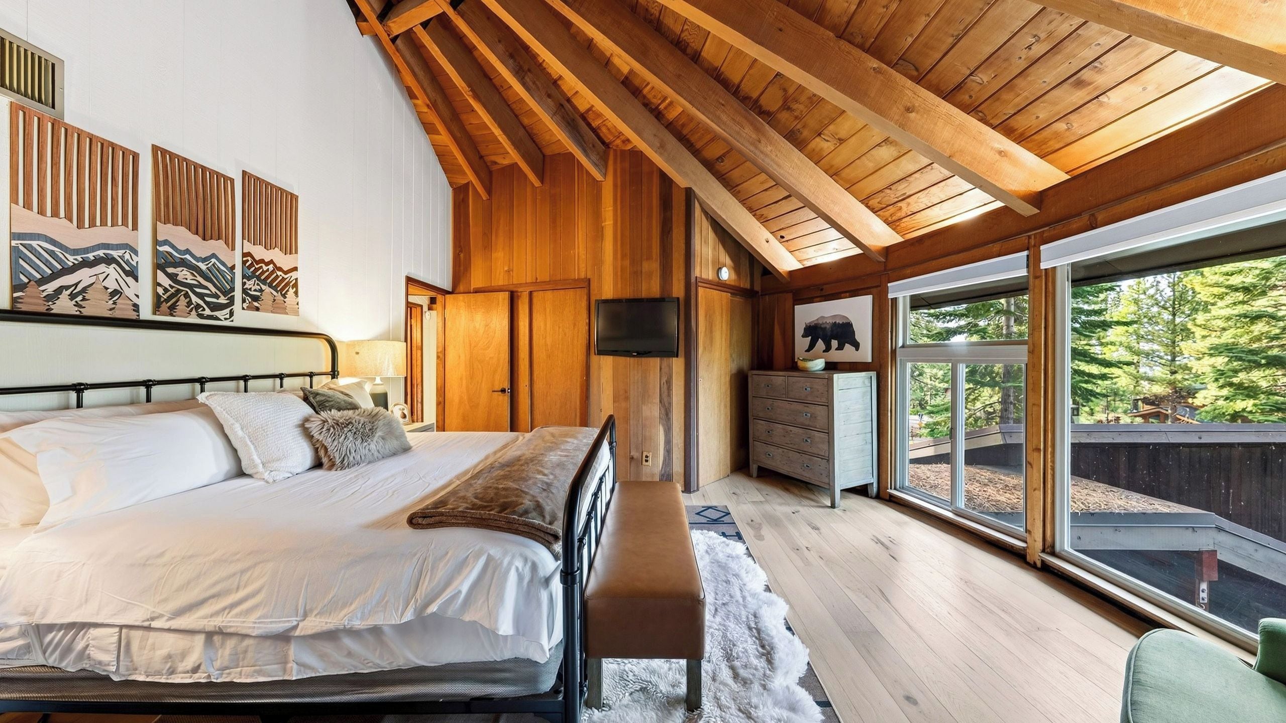 A bedroom with a large bed, wooden ceiling beams, a dresser, and a window with a view of trees.