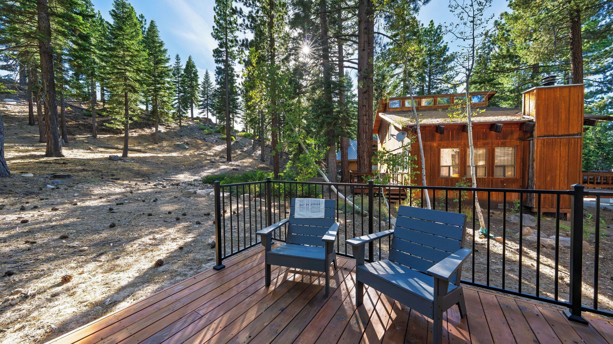 Eine Holzterrasse mit zwei Stühlen und einem Metallgeländer, umgeben von Bäumen und einer Holzhütte.