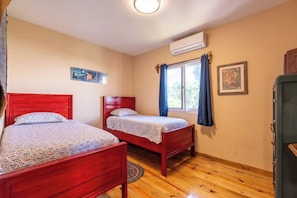 Ein Schlafzimmer mit zwei Holzbetten, einem fenster mit Vorhängen, einer Klimaanlage und einem gerahmten Bild an der Wand.