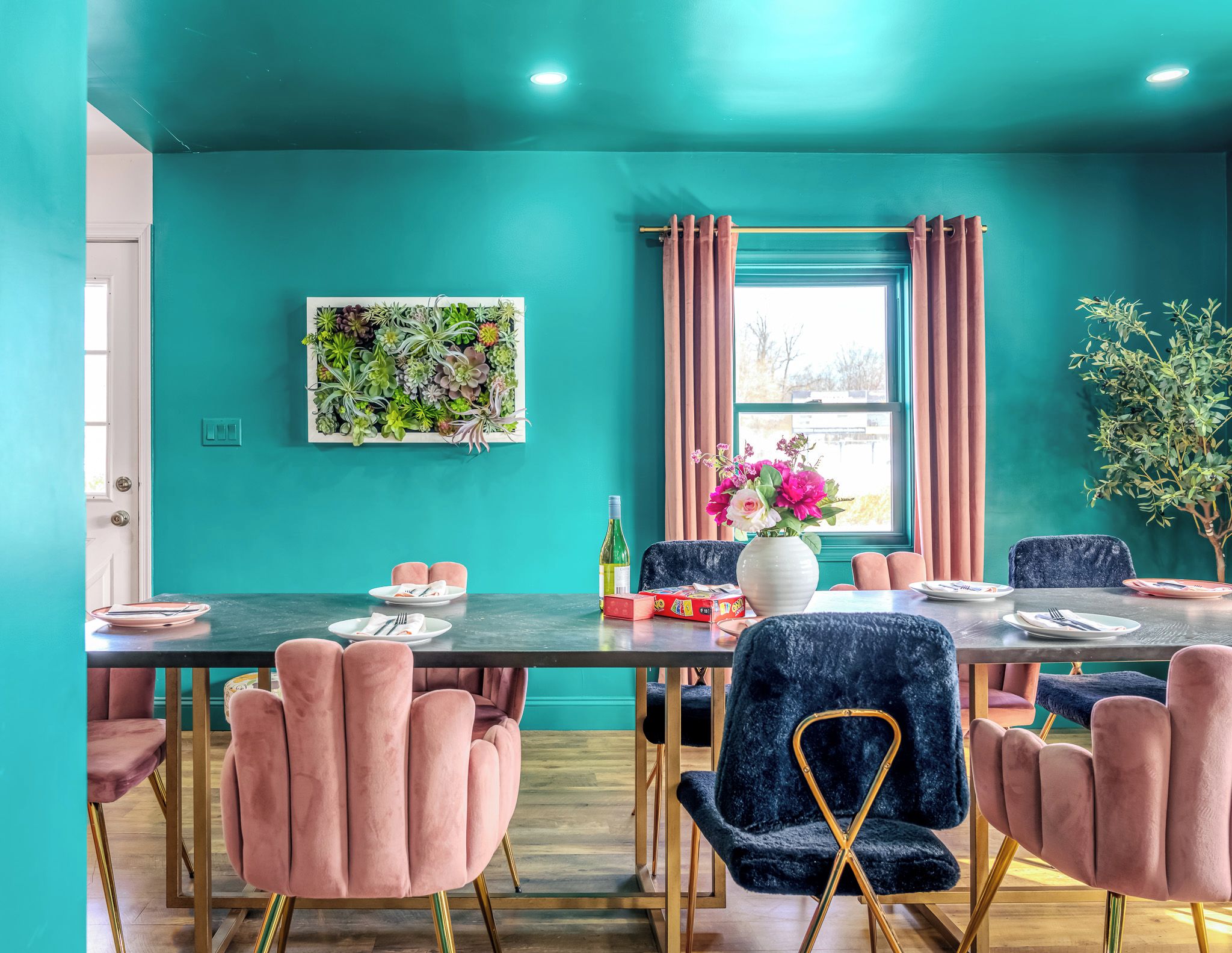 A teal-colored room with a dining table set for four, surrounded by plush chairs with gold legs.