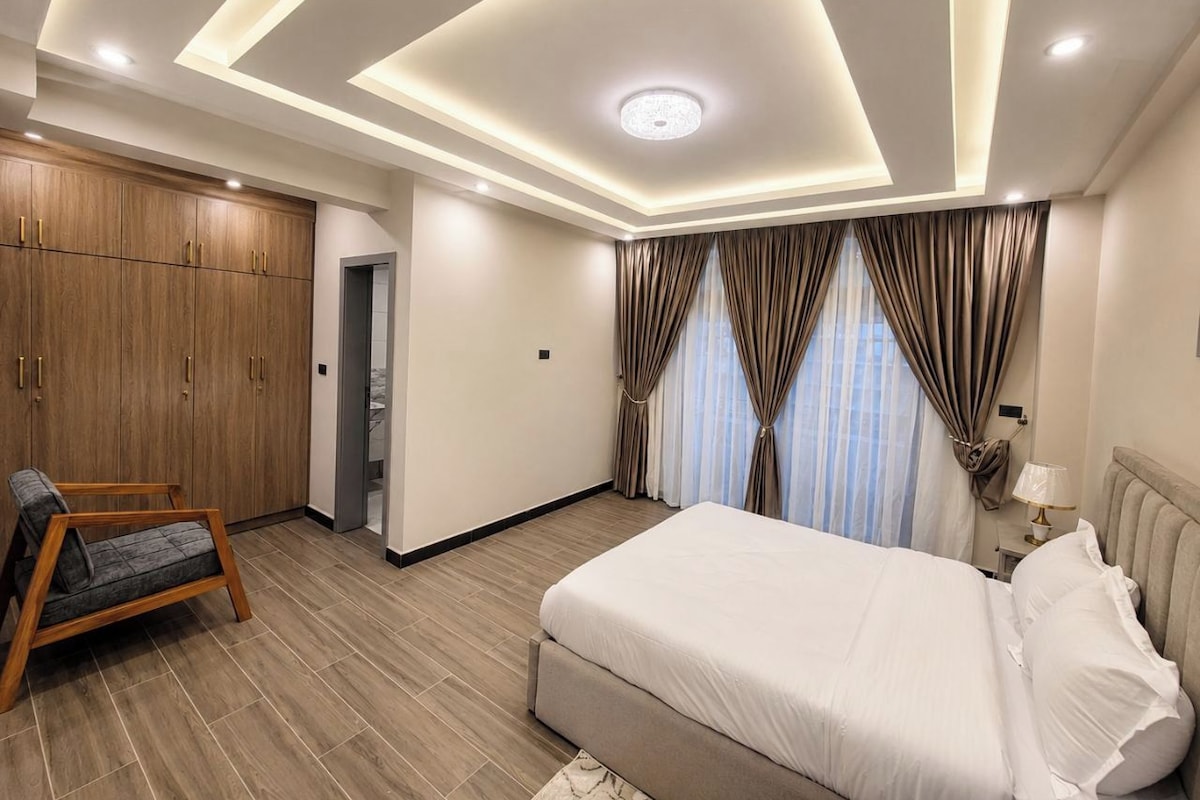 Quarto de hotel com uma cama grande, guarda-roupa, área de estar e teto com iluminação embutida.