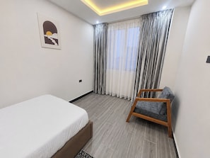 Quarto de hotel com cama, uma cadeira e uma janela grande com cortinas.