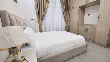 Quarto moderno com cama, mesa de cabeceira, luminária, cortinas e armário embutido.