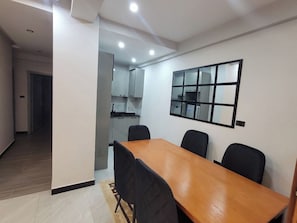 Uma área de refeições com mesa de madeira e cadeiras pretas, cozinha ao fundo e um espelho fixado na parede.