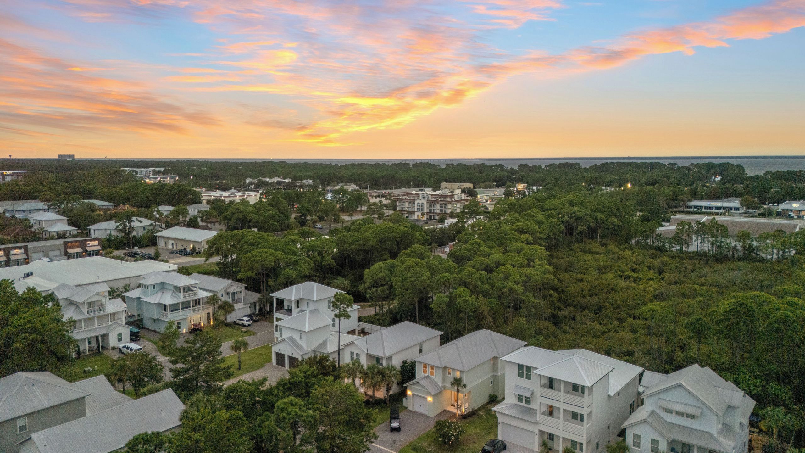 A residential area with multiple two-story houses, surrounded by dense greenery and a clear view of the ocean in the distance.