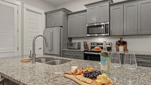 A modern kitchen with grey cabinets, stainless steel appliances, granite countertops, and a variety of food items. - Maré 33 by Avantstay 6-seat Golf Cart + Arcade + Bunk Room Beach Walk + Pool (Miramar Beach)