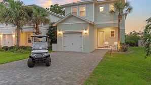 A residential property with a paved driveway, a golf cart, and a house with a garage and a porch with chairs. - Maré 33 by Avantstay 6-seat Golf Cart + Arcade + Bunk Room Beach Walk + Pool (Miramar Beach)
