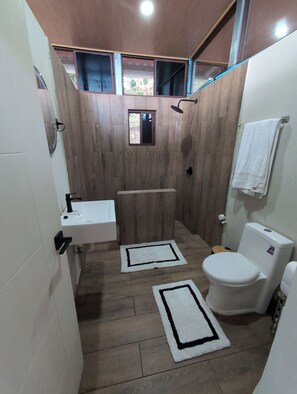 A bathroom with a wooden floor, a white sink, a toilet, and a shower. There are two rugs on the floor.