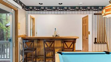 A pool table in a room with a bar area, two chairs, and a door leading outside.