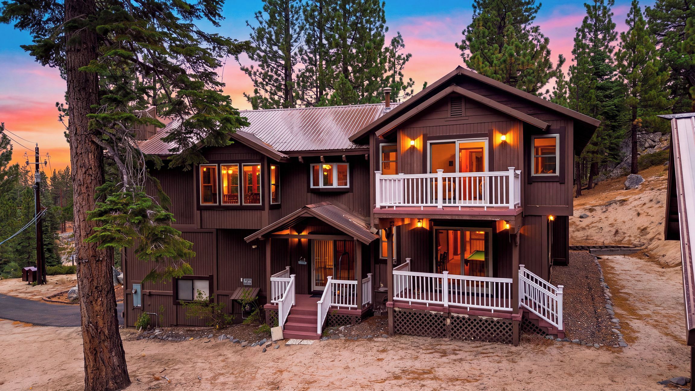 A two-story wooden cabin with a balcony, surrounded by trees and a dirt driveway.