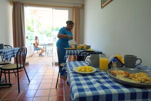 Un comedor con manteles a cuadros, una persona sirviendo comida y alimentos para el desayuno, como huevos y fruta.