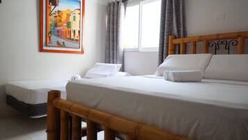 Habitación de hotel con una cama grande, otra más pequeña, un cabecero de madera, una ventana con cortinas y un cuadro en la pared.