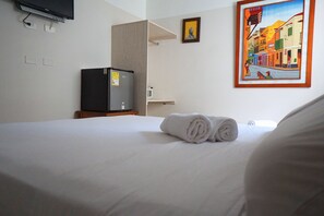 Habitación de hotel con cama, televisión, refrigerador pequeño y un cuadro en la pared.