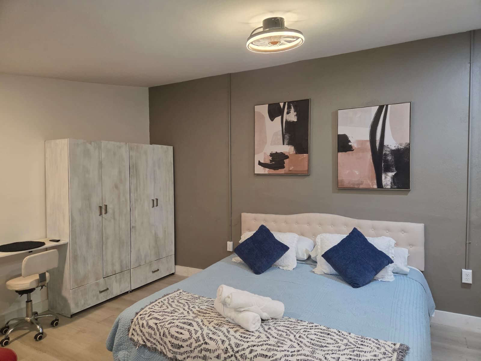 A bedroom with a bed, two pillows, a bedside table, a chair, a wardrobe, and two abstract paintings on the wall.