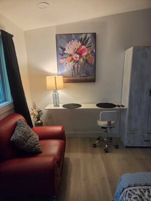 Een hotelkamer met een rode leren bank, een bureau met een lamp, een schilderij van bloemen en een kledingkast.
