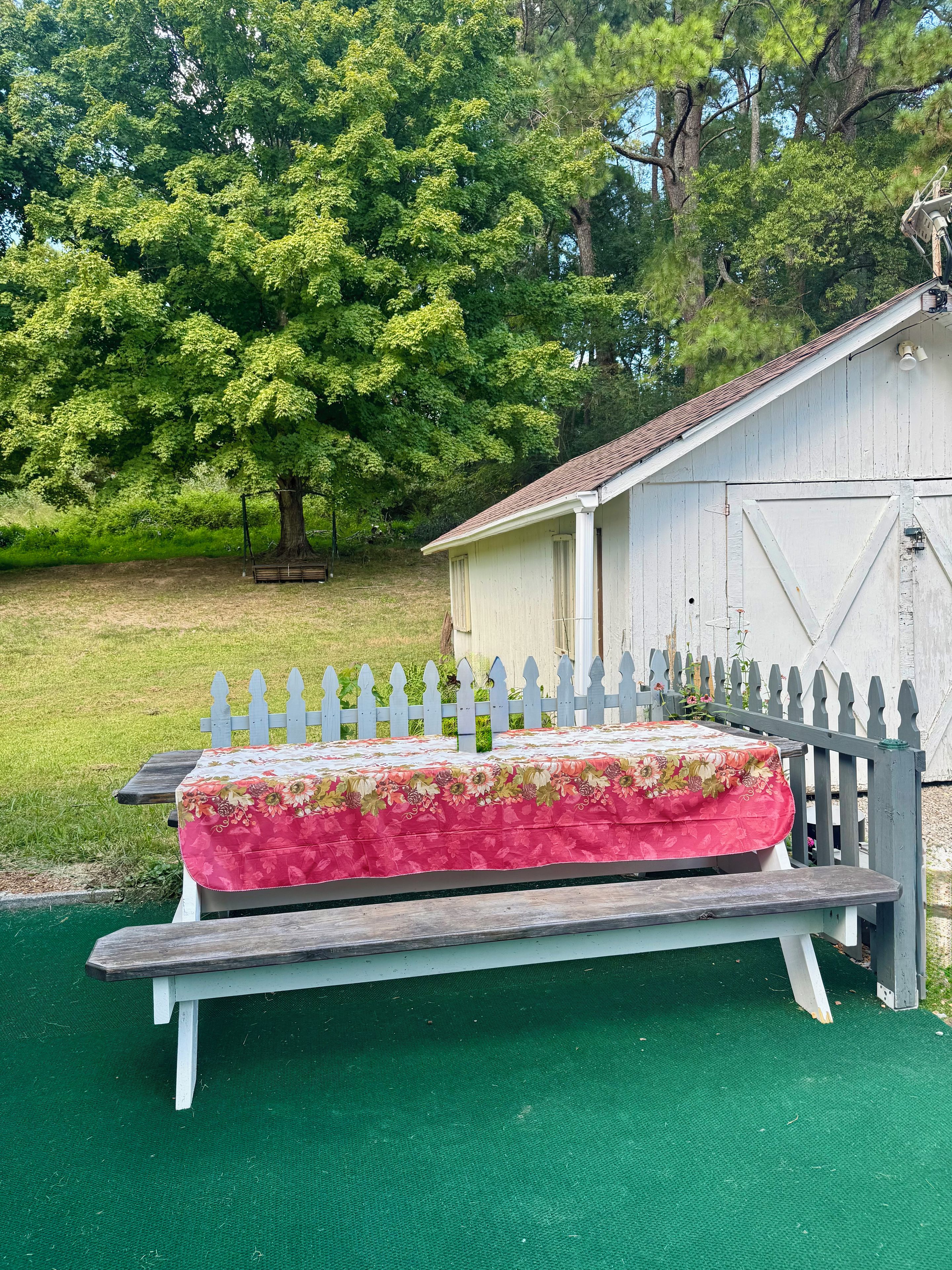 Ein Bett mit floralem Überwurf, eine Holzbank und ein weißer Zaun in einem Garten mit Baum und Gartenhaus.