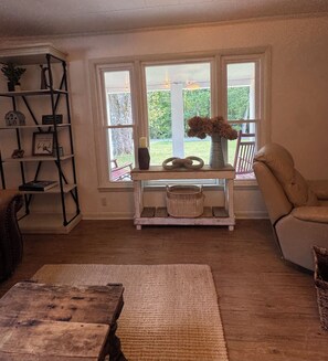 Una habitación con piso de madera, un sofá, una silla, una mesita con un jarrón y una estantería.
