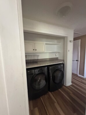 Un área de lavandería compacta con dos lavadoras de carga frontal, una encimera con una cesta metálica y un armario blanco arriba.