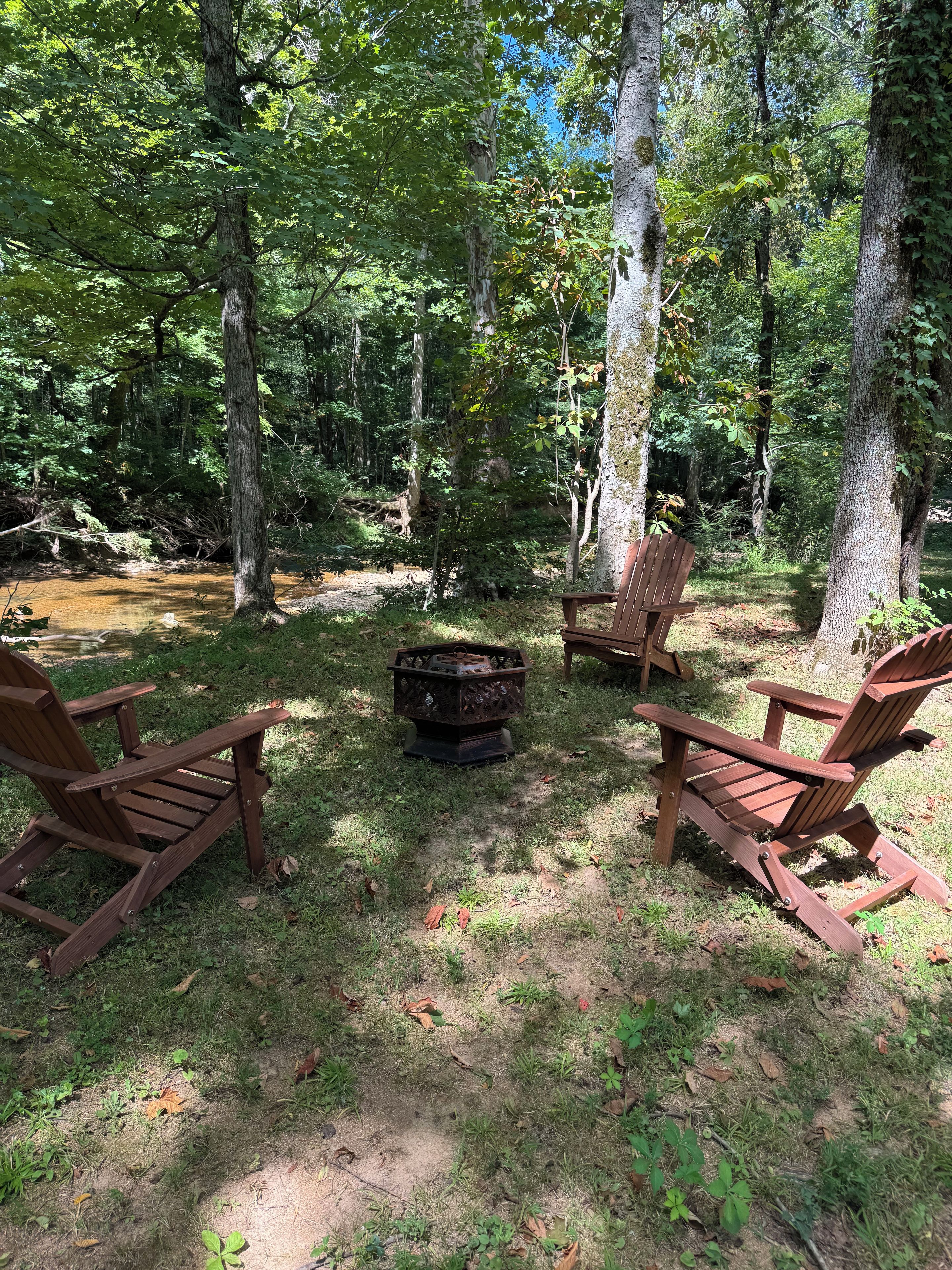 A forested area with two wooden Adirondack chairs and a fire pit.