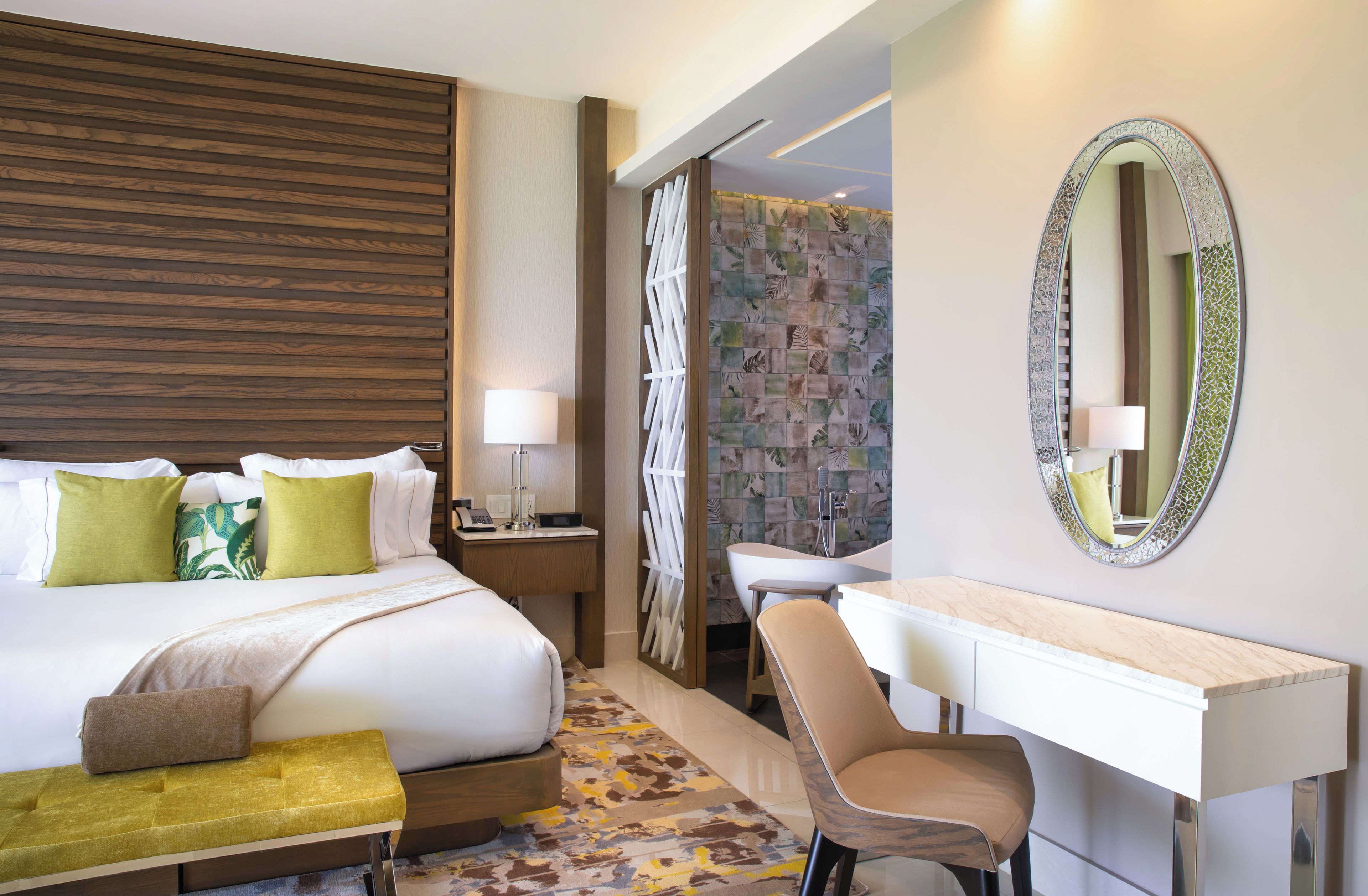 A hotel room with a large bed, a desk, a chair, a vanity, and a shower area.