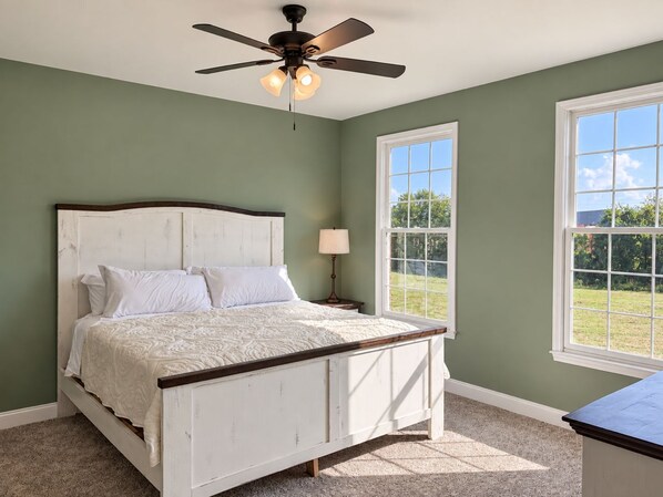 A bedroom with a bed, a ceiling fan, a nightstand, a lamp, and a window with a view of the outdoors.