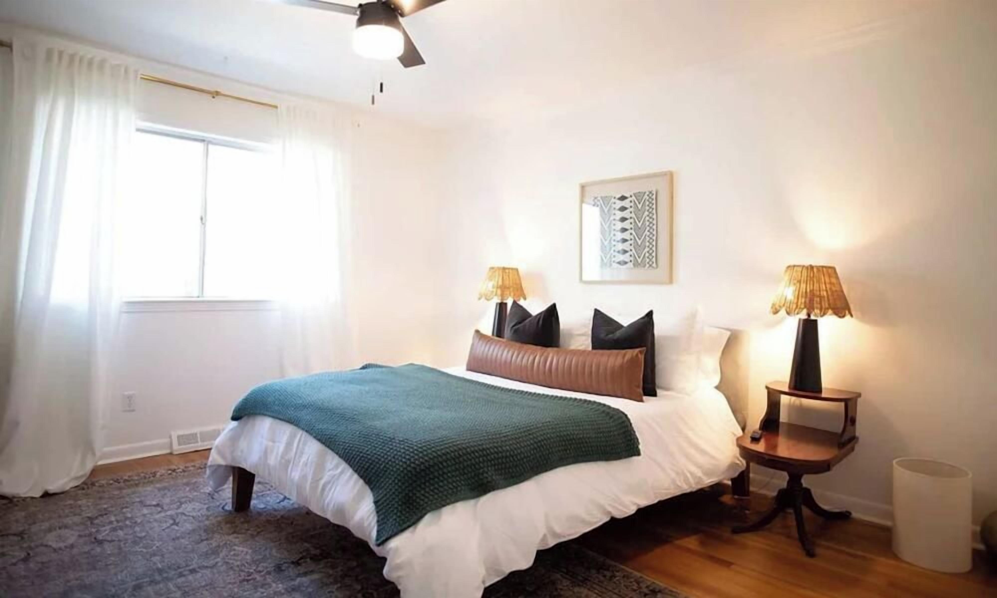 Un dormitorio con una cama, dos mesitas de noche con lámparas, una ventana con cortinas y una obra de arte colgada en la pared.