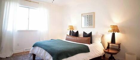 Un dormitorio con una cama, dos mesitas de noche con lámparas, una ventana con cortinas y una obra de arte colgada en la pared.