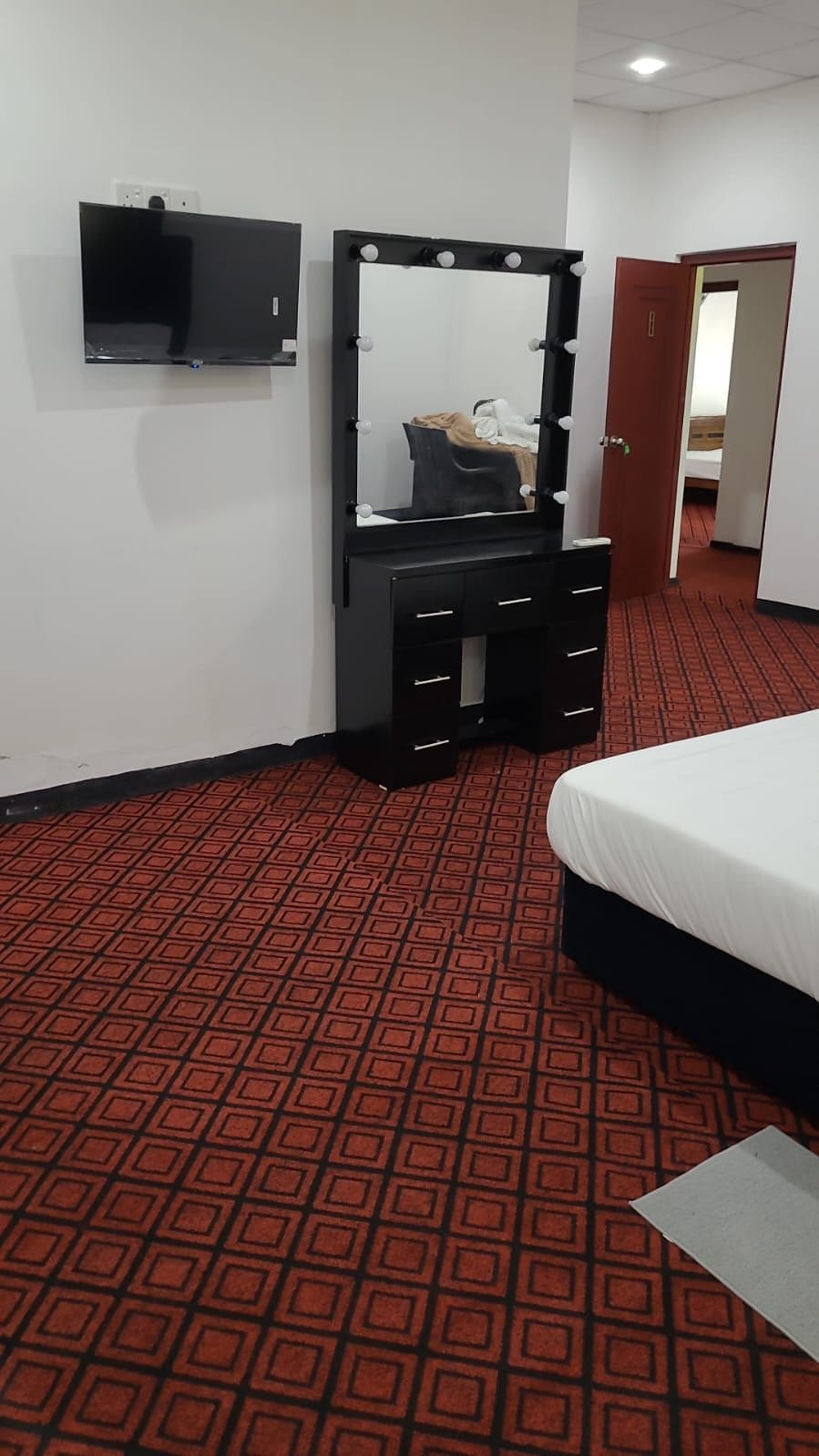 Habitación de hotel con cama, tocador con espejo e iluminación, televisor de pantalla plana y alfombra estampada.