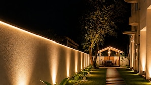 A nighttime outdoor pathway with a well-lit wall, a tree, and a covered seating area. - Amore Beach Resort (Balapitiya)