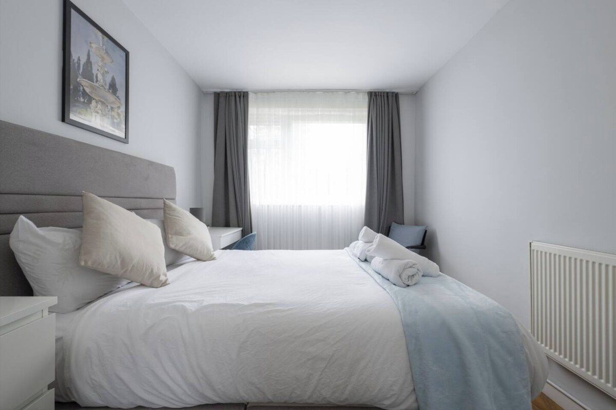 A neatly made bed with white bedding, a grey headboard, and a white radiator.