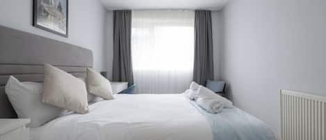 A neatly made bed with white bedding, a grey headboard, and a white radiator.