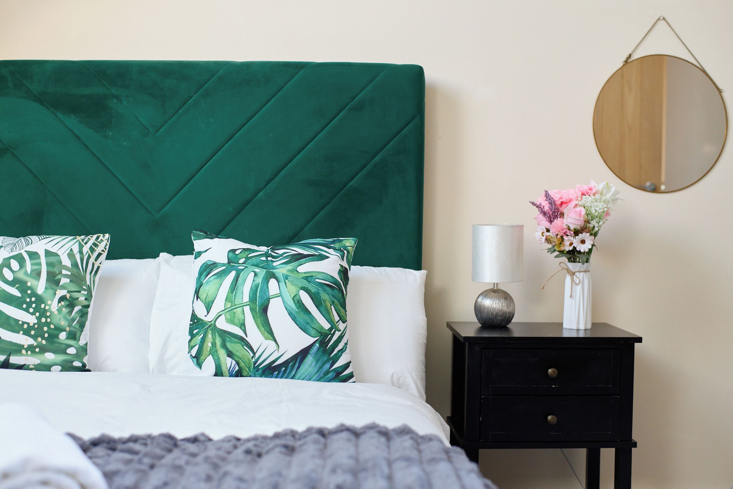 Ein Bett mit einem grünen, gepolsterten Kopfteil, weißen Kissen, einer grauen Decke, einem Nachttisch mit Lampe und einer Vase mit Blumen.