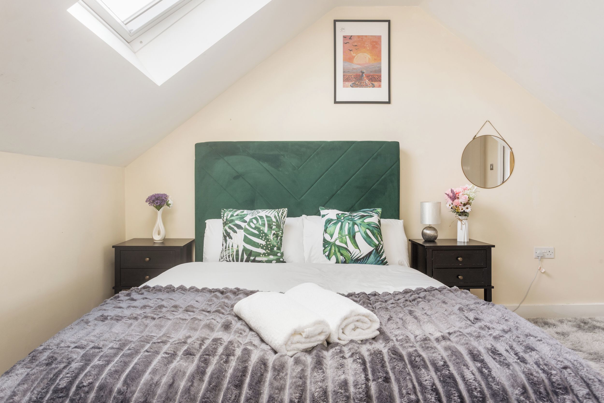 Une chambre avec un plafond en pente, un puits de lumière, un lit doté d’une tête de lit recouverte d’un tissu vert, deux tables de chevet, un miroir rond et un tableau encadré accroché au mur.