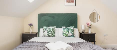 Un dormitorio con techo inclinado, tragaluz, cama con cabecera tapizada en verde, dos mesitas de noche, un espejo redondo y un cuadro en la pared.