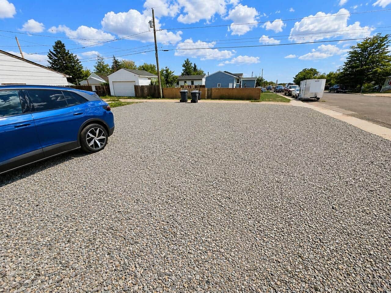 A residential driveway with a blue car parked on the left, a gravel surface, and houses in the background.